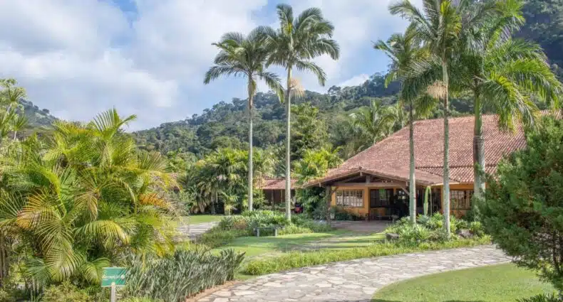 Um jardim tropical com palmeiras altas, um caminho de pedras e um edifício com telhado de telhas vermelhas, tendo como pano de fundo colinas verdes. Esse é o Bomtempo Itaipava, um dos hotéis fazenda no Rio de Janeiro.