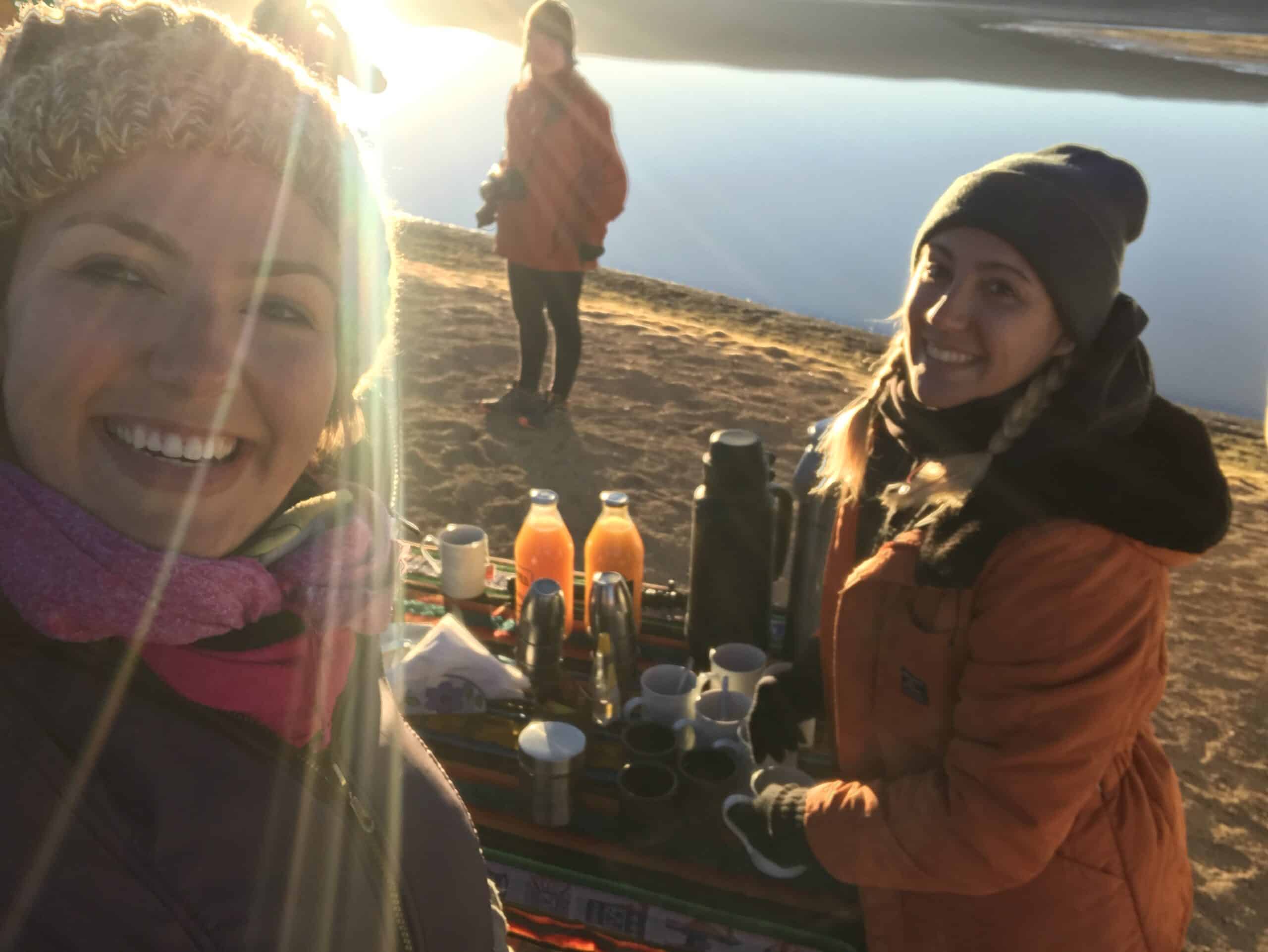 Três pessoas ao ar livre perto de um corpo d'água. Duas estão sorrindo para a câmera, com mesas segurando bebidas e garrafas térmicas na frente delas.