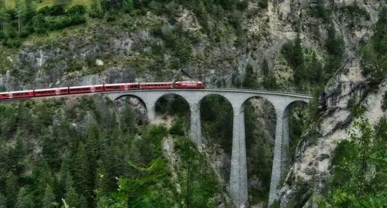 Um trem vermelho cruza o Viaduto Landwasser, uma ponte de pedra com arcos altos, situada em uma área montanhosa e florestal, ilustrando a capa do post como viajar de trem pela Suíça.