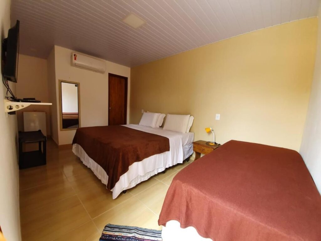 Quarto da Casa do Maneco com uma cama de casal e uma cama de solteiro, TV montada na parede, espelho, ar condicionado e uma pequena mesa com luminária.
