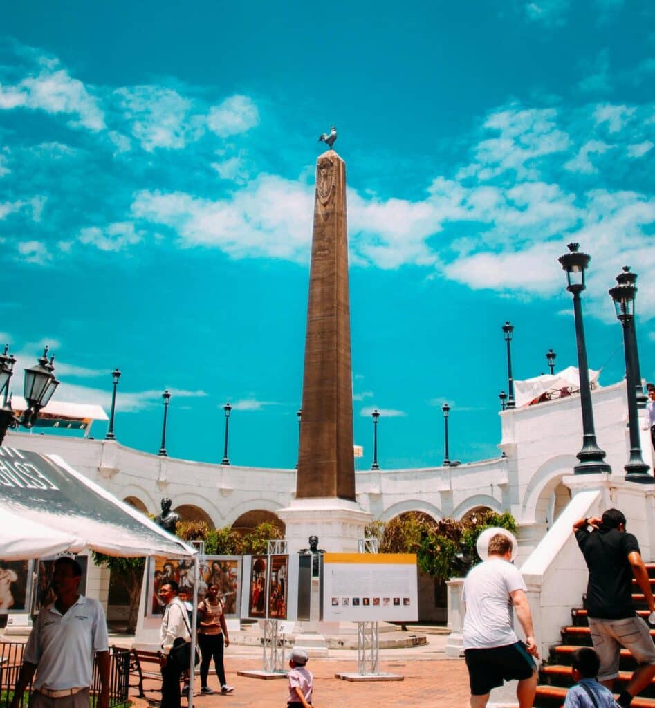 Pessoas caminham perto de um alto obelisco em Casco Viejo, cercado por arcos e postes de luz sob um céu azul claro. A foto ilustra o post de o que fazer na Cidade do Panamá.