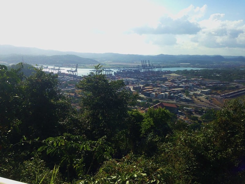 Vista do topo de Cerro Ancon. A imagem mostra um porto movimentado com guindastes, contêineres e vegetação ao redor sob um céu parcialmente nublado.