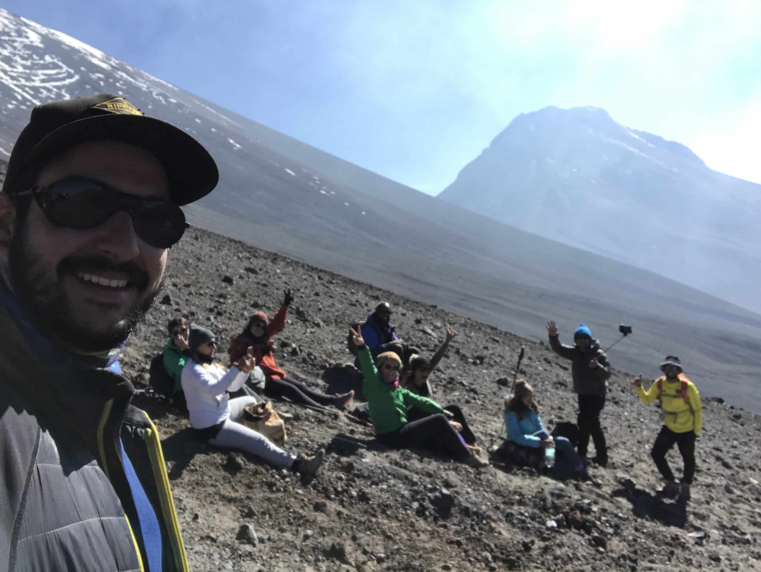 Grupo de pessoas sentadas em uma encosta rochosa com uma montanha nevada ao fundo, uma pessoa tirando uma selfie, outras acenando, na trilha do vulcão Láscar no deserto do Atacama.