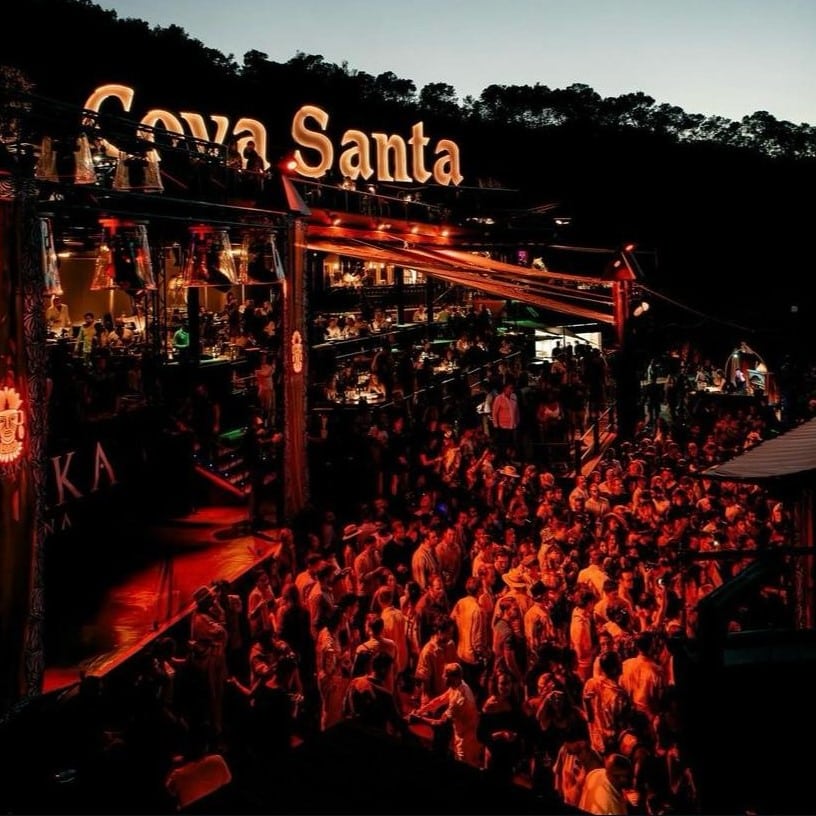 Um movimentado local ao ar livre, cheio de pessoas sob o céu noturno, iluminado por uma luz vermelha quente, com uma placa "Cova Santa" acima. Essa é uma das melhores festas em Ibiza.