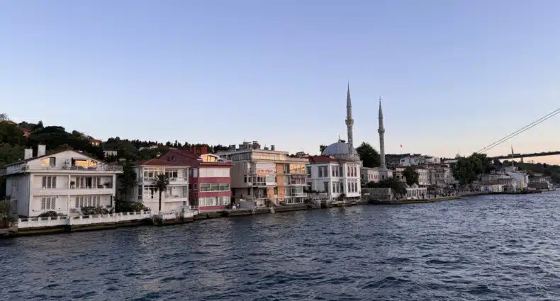 Edifícios à beira-mar ao longo de uma costa com uma mesquita com dois minaretes ao fundo sob um céu claro, um cruzeiro pelo Bósforo.
