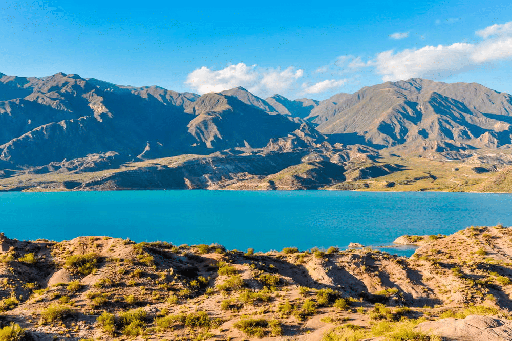 Vista do Dique de Potrerillos, um lago azul claro cercado por colinas rochosas e montanhas distantes sob um céu azul brilhante com nuvens dispersas.