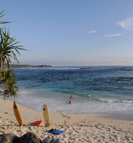 Praia Dream Beach em Nusa Lembongan de areia branca com duas pranchas de surfe na areia, pessoas na água e uma árvore à esquerda, sob um céu azul claro.