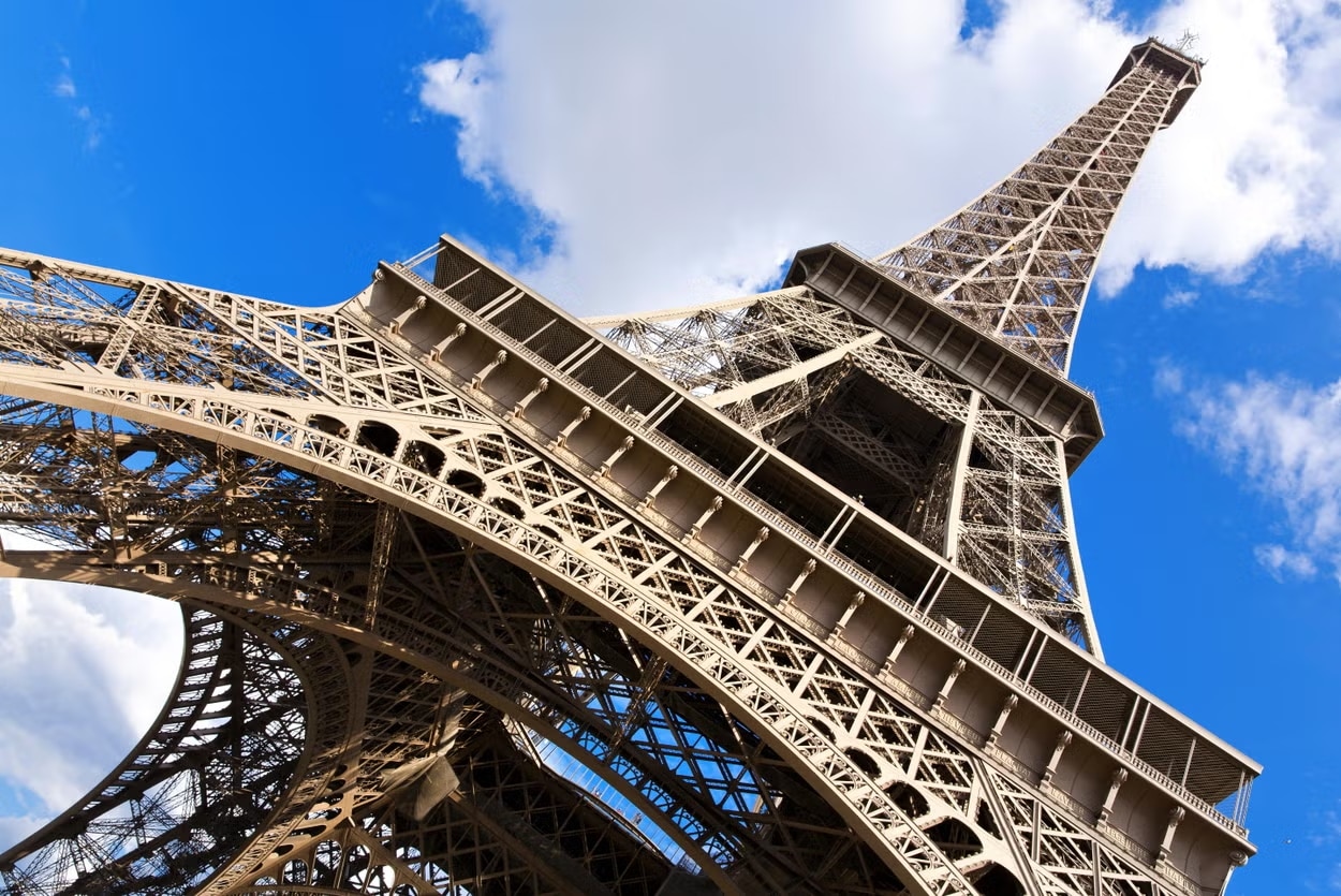 Vista de ângulo baixo da Torre Eiffel contra um fundo de céu azul e nuvens brancas para ilustrar o post sobre pontos turísticos de Paris.