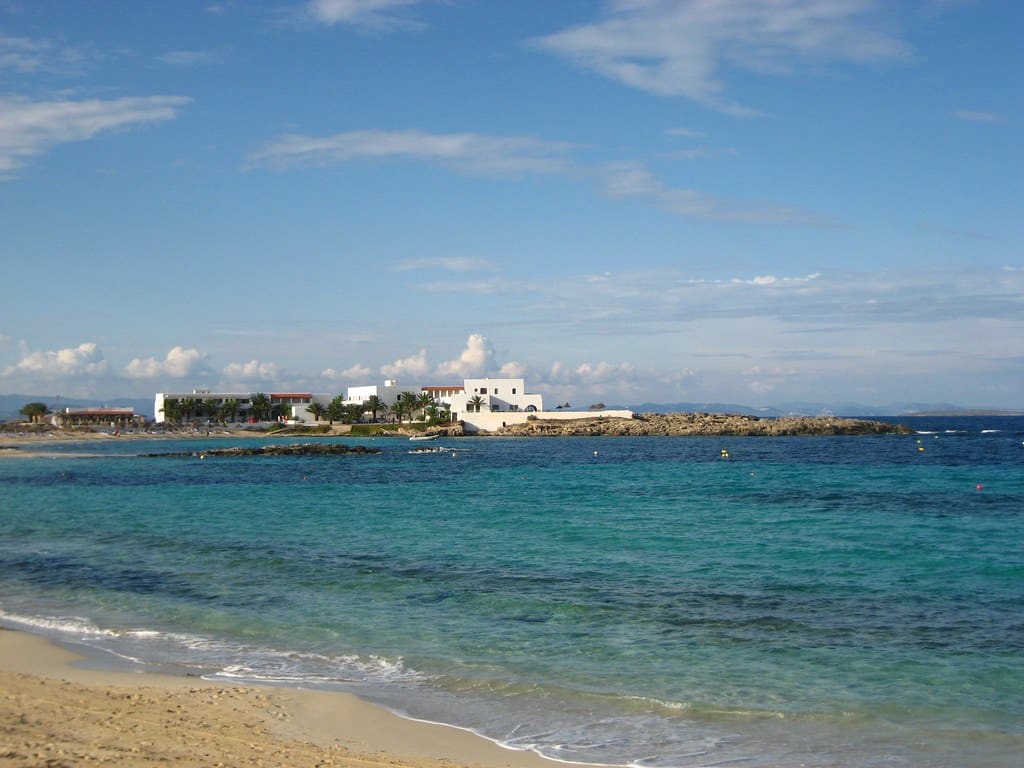 Vista de uma praia com areia fina, água azul-turquesa e um edifício branco ao longe sob um céu parcialmente nublado no vilarejo de Es Pujols, uma das opções de o que fazer em Formentera.
