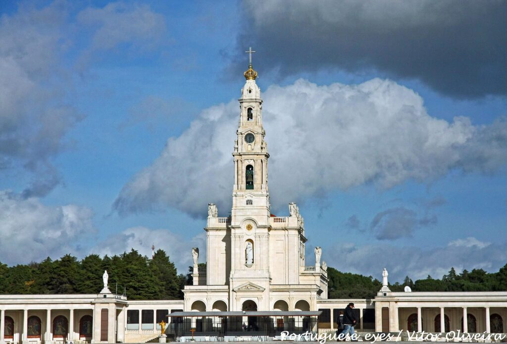 A Basílica de Nossa Senhora do Rosário, com uma torre alta e uma cruz, em contraste com um céu azul com nuvens, cercada por árvores.