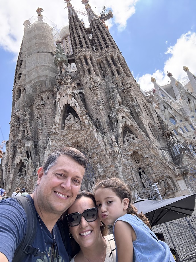 Uma família de três pessoas posa em frente à Sagrada Família em Barcelona em um dia ensolarado, com as torres da basílica ao fundo.