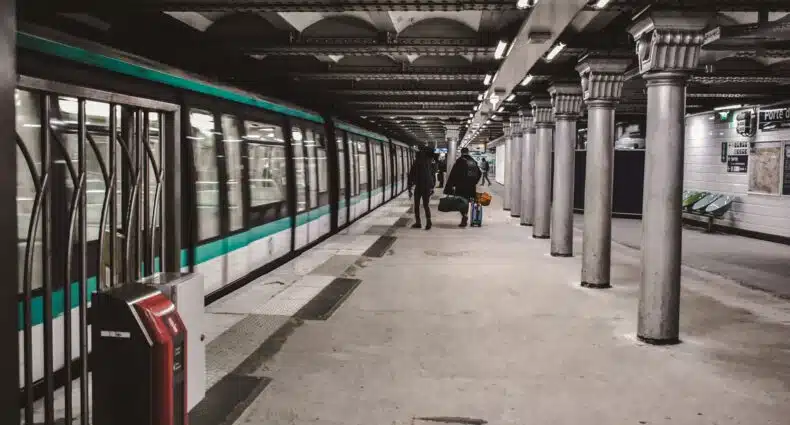 Um trem do metrô de Paris está chegando a uma estação subterrânea com pessoas em pé na plataforma ao lado de colunas de metal. A imagem ilustra o post sobre transportes em Paris.