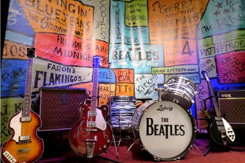 Uma coleção de guitarras, amplificadores e uma bateria com o logotipo "The Beatles" em frente a uma parede colorida com nomes de bandas no Museu dos Beatles, uma das opções de o que fazer em Canela.