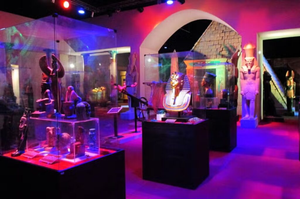 Exposição do Museu Egípcio de Canela com vários artefatos egípcios antigos em vitrines de vidro, iluminação colorida e estátuas ao longo das paredes.