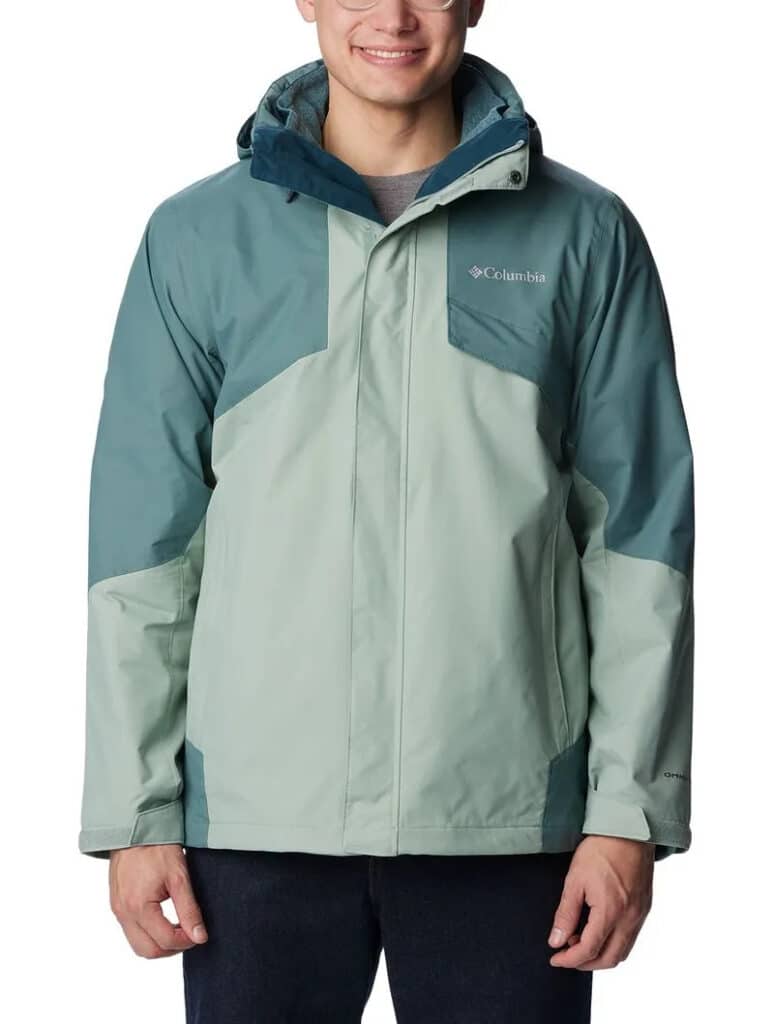 Uma pessoa vestindo uma jaqueta Columbia verde bicolor com capuz, em pé, contra um fundo branco.