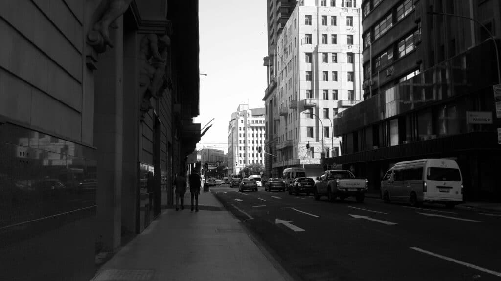 Imagem em preto e branco de uma rua urbana com carros, uma van e pessoas caminhando na calçada ao lado de edifícios altos em Joanesburgo.