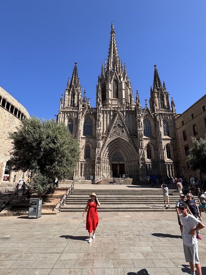 Uma mulher de vestido vermelho caminha em frente a uma grande catedral de estilo gótico em um dia claro. Essa é a Catedral de Barcelona, uma das opções de o que fazer em Barcelona.