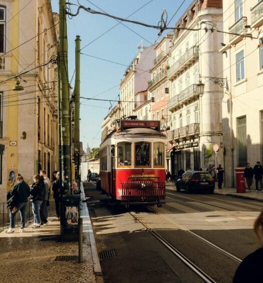 Um bonde vermelho em Lisboa viaja por uma rua repleta de prédios e pedestres sob um céu azul claro. Representa quando ir para Portugal.