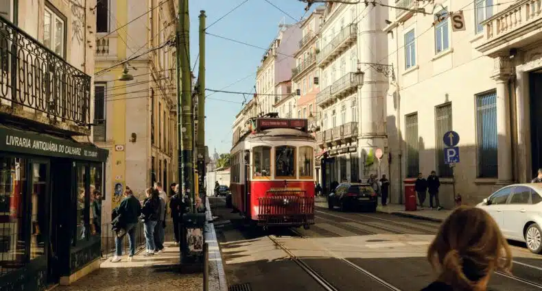 Um bonde vermelho em Lisboa viaja por uma rua repleta de prédios e pedestres sob um céu azul claro. Representa quando ir para Portugal.