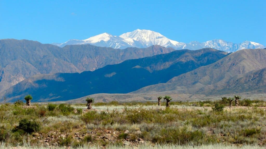 Paisagem com montanhas cobertas de neve ao fundo, terreno árido e gramado em primeiro plano e céu azul claro acima.