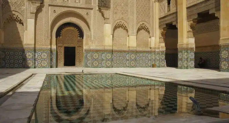 Pátio da Madrassa Ben Youssef primorosamente decorado com uma porta em arco e um espelho d'água, cercado por paredes de azulejos estampados. Imagem para ilustrar post sobre o que fazer em Marrakech.