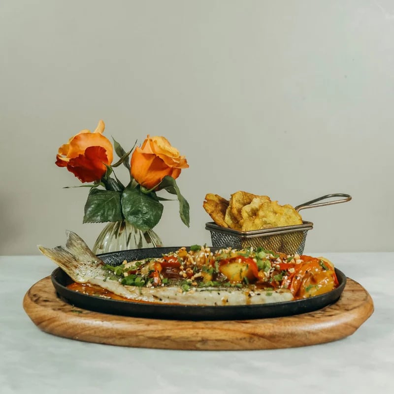 Prato do Mahalo Cocina y Jardin com peixe grelhado com legumes. Ele é servido em uma bandeja de madeira ao lado de uma cesta de batatas fritas e um vaso com rosas laranja.