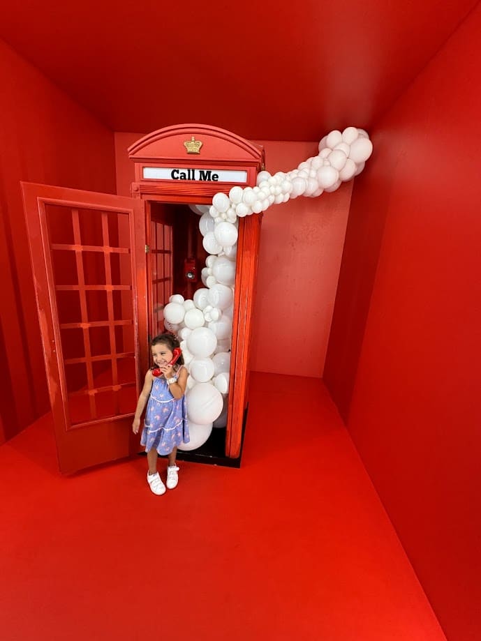 Uma criança de vestido azul está dentro de uma cabine telefônica vermelha decorada com balões brancos em uma sala vermelha no Museu Nacional de Arte da Catalunha.