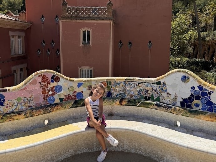 Criança sentada em um banco de mosaico colorido com um edifício ornamentado e árvores ao fundo no Parque Güell.