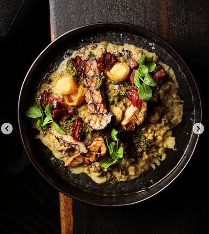 Uma tigela de risoto cremoso de cogumelos coberto com legumes grelhados, verduras frescas e ervas em uma mesa de madeira escura no Marble Restaurant.