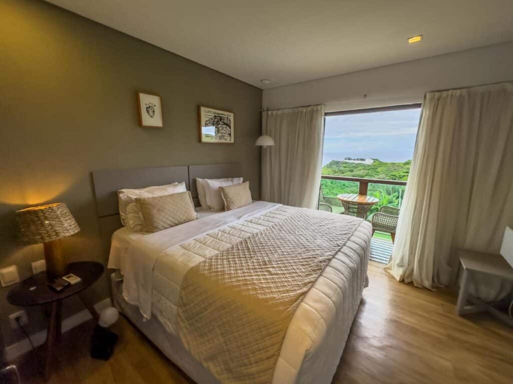 Quarto aconchegante no Miragem Noronha com cama de casal, tons neutros e sacada com vista para a vegetação exuberante. Uma luminária e arte emoldurada adornam o quarto.