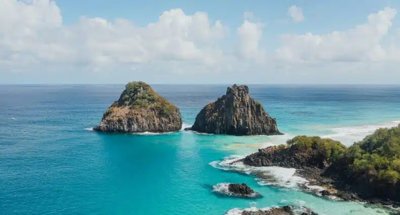 Duas ilhas rochosas cercadas por água turquesa do oceano sob um céu azul com nuvens. A vegetação é visível nas ilhas. É o Morro Dois Irmãos em Noronha.