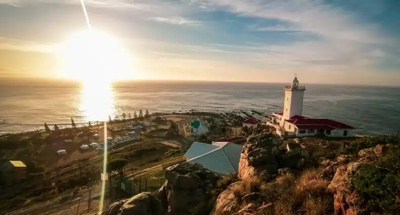 Pôr do sol em Mossel Bay sobre uma paisagem costeira com um farol, edifícios e terreno rochoso, com vista para o oceano. Representa na Rota do Jardim na África do Sul.