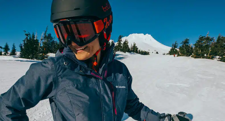 Uma pessoa usando equipamento de esqui, jaqueta da Columbia e óculos sorri enquanto está em uma encosta de montanha nevada com árvores e um pico ao fundo. Essa imagem ilustra o post sobre jaqueta para neve.