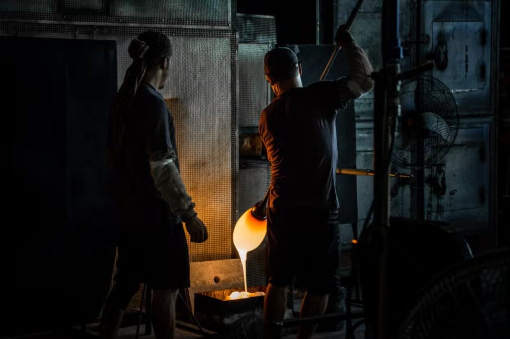 Dois sopradores de vidro trabalham com vidro derretido em uma oficina escura, iluminados pelo material brilhante enquanto o moldam cuidadosamente no Museu Cristais de Gramado