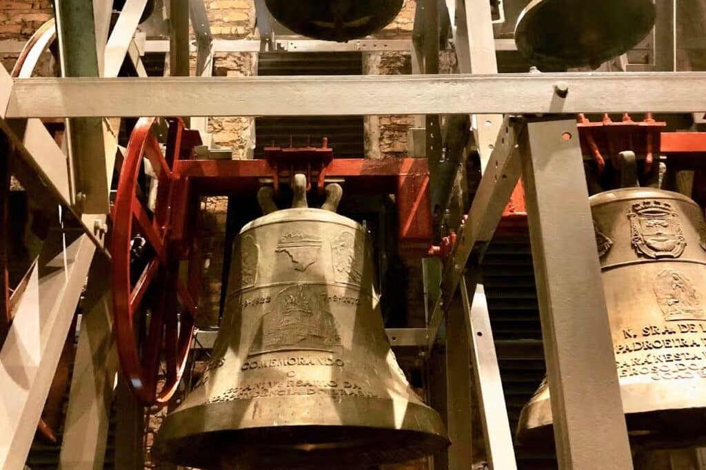 Um close-up de grandes sinos de metal montados em uma estrutura de torre sineira, com gravuras complexas e componentes de metal vermelho no Museu dos Sinos da Catedral em Canela.