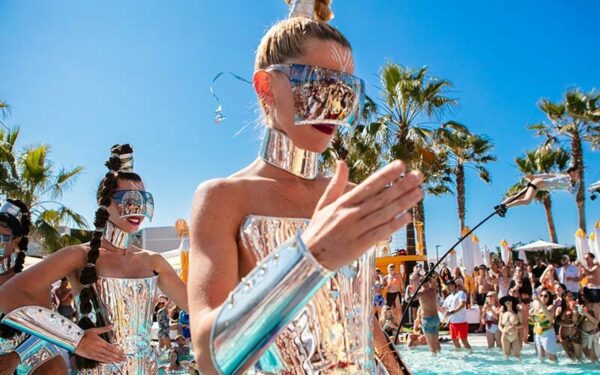 Artistas em trajes e viseiras metálicas caminham entre uma multidão em uma piscina sob um céu azul claro com palmeiras ao fundo no O Beach Ibiza, uma das festas em Ibiza.