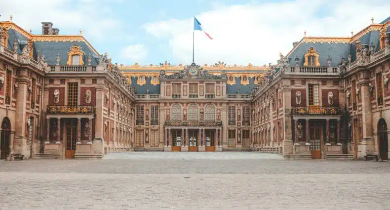 O pátio do Palácio de Versalhes com um edifício central, detalhes dourados ornamentados e uma bandeira francesa no topo, sob um céu parcialmente nublado.