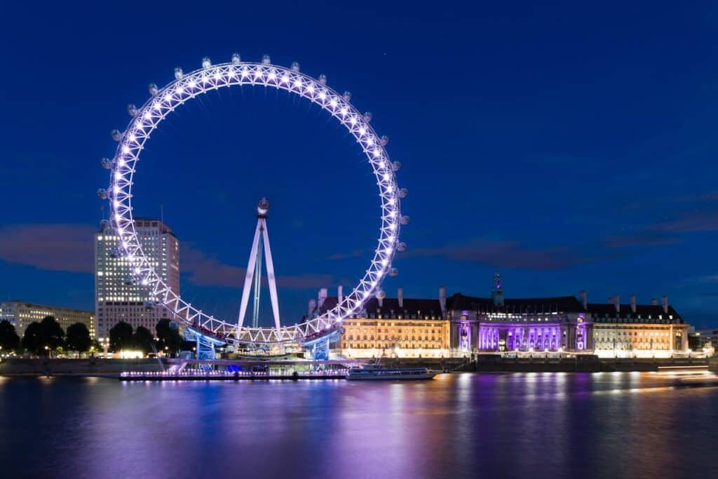 Roda gigante iluminada e edifícios perto de um rio à noite, com reflexos na água e um céu azul escuro e claro. A imagem ilustra o post sobre roteiro para Londres.