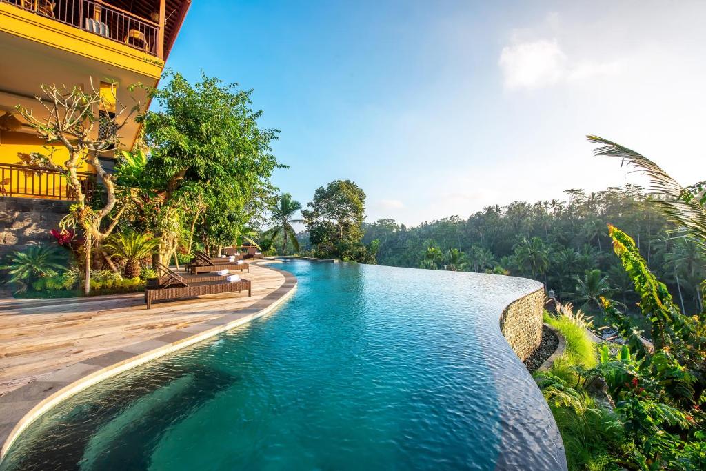 Piscina de borda infinita do GK Bali Resort com vista para uma paisagem verdejante e céu azul claro, ao lado de um prédio amarelo e cercada por árvores.