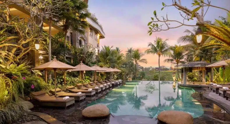 Uma luxuosa área à beira da piscina com espreguiçadeiras, guarda-sóis e plantas tropicais ao pôr do sol, cercada por um exuberante jardim, no hotel Kuwarasan a Pranama, uma das dicas de onde ficar em Bali.