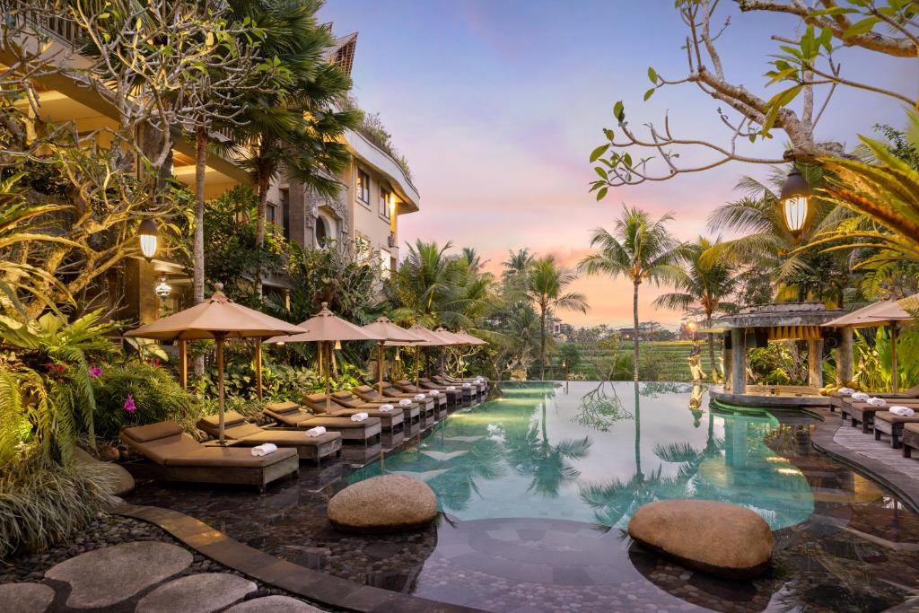 Uma luxuosa área do Kuwarasan A Pramana Experience à beira da piscina com espreguiçadeiras, guarda-sóis e plantas tropicais ao pôr do sol, cercada por um exuberante jardim.