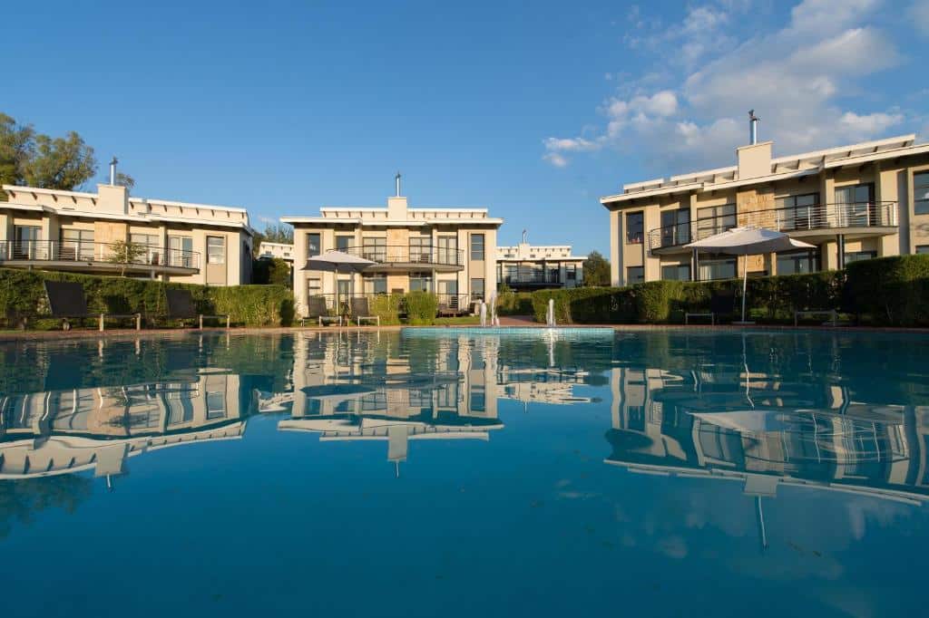 Moradias modernas do The Fairway Hotel, Spa & Golf Resort
com janelas grandes são refletidas em uma piscina azul-clara. Guarda-sóis e vegetação estão ao fundo. Representa Joanesburgo.