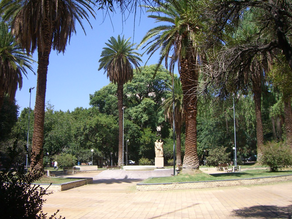 Plaza Chile com palmeiras altas, uma estátua de pedra no centro, caminhos de tijolos e bancos sob um céu azul claro. Foto para ilustrar post sobre o que fazer em Mendoza.