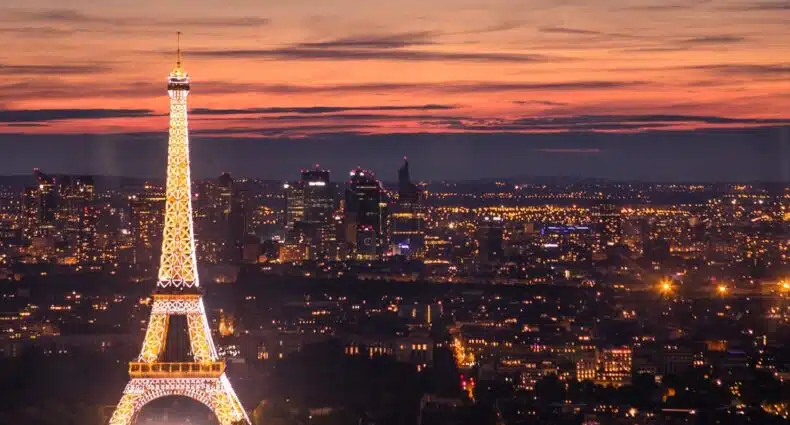 A Torre Eiffel é iluminada contra um pôr do sol vibrante, com vista para as luzes da cidade de Paris ao fundo. A imagem ilustra o post sobre onde assistir o pôr do sol em Paris.