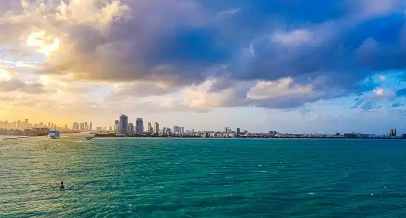Horizonte da cidade de Miami com edifícios altos sob um céu parcialmente nublado, com vista para um vasto corpo de água azul-turquesa ao pôr do sol. Representa Miami.