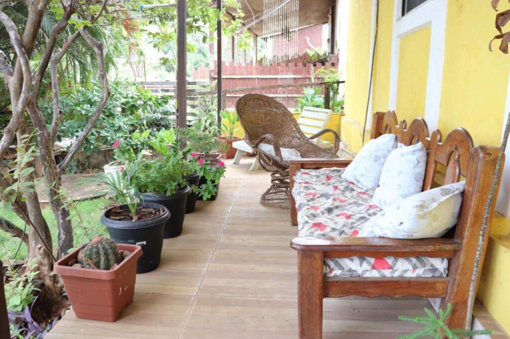 Uma varanda da Pousada Água Viva com cadeiras de madeira, almofadas estampadas, uma cadeira de vime, vasos de plantas e uma parede amarela ao fundo.