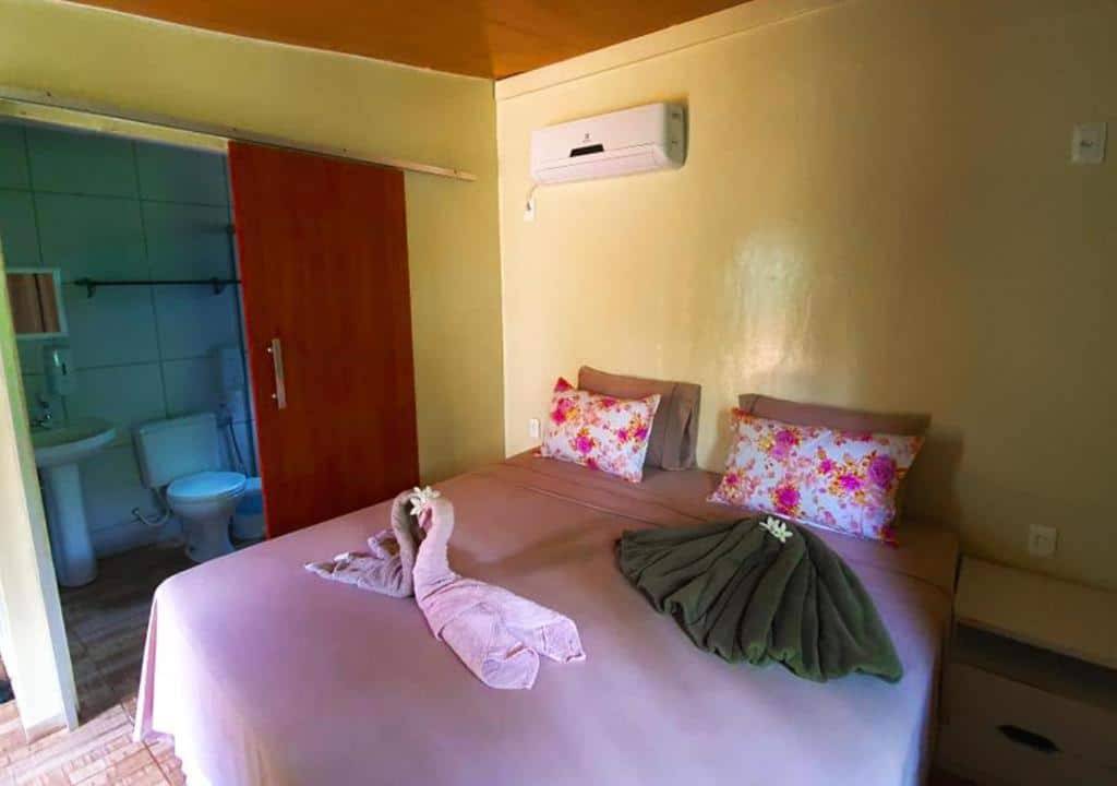 Um quarto aconchegante com uma cama de casal, travesseiros florais, toalhas dobradas, ar condicionado e um banheiro anexo com vaso sanitário e pia. Representa o post de Como economizar em Fernando de Noronha.