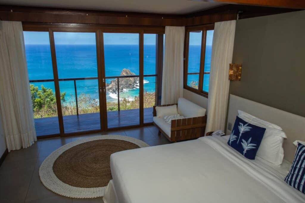 Um quarto da Pousada Ecológica Akanã com uma cama de casal, tapete redondo e sofá, com grandes janelas com vista para o oceano e o litoral rochoso.