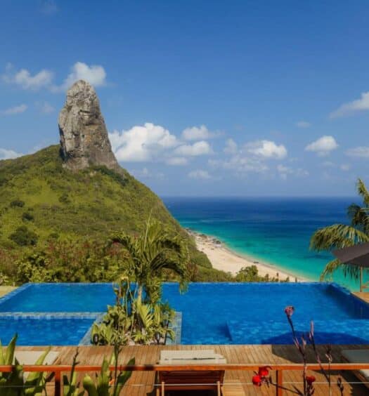 Piscina de borda infinita da Pousada Ecológica Akanã com vista para a montanha e a praia sob um céu azul claro, cercada por vegetação exuberante e espreguiçadeiras. Imagem para ilustrar post sobre onde ficar em Fernando de Noronha.