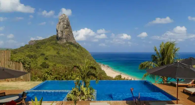 Piscina de borda infinita da Pousada Ecológica Akanã com vista para a montanha e a praia sob um céu azul claro, cercada por vegetação exuberante e espreguiçadeiras. Imagem para ilustrar post sobre onde ficar em Fernando de Noronha.